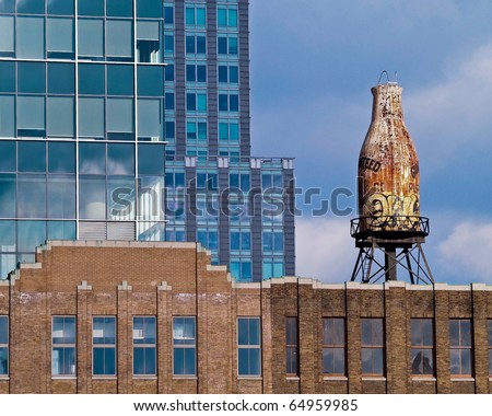 a city scape with three buildings of different styles. a giant rusted bottle sits on top of one of the buildings.