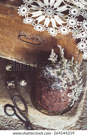 Homemade Chocolate Cake with Coconut Flakes and Dried Flower Decoration on a Rough Concrete Floor. Vintage Chest with a Lace Cover in the Background