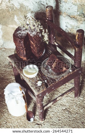 Homemade Chocolate Cake with Coconut Flakes and Dried Flower Decoration Next to a Bowl of Yogurt on a Small Vintage Chair. Glass Jar with a Wooden Spoon on the Floor. Rough Concrete Background