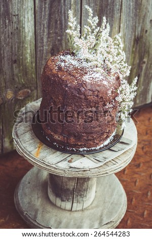Homemade Chocolate Cake with Coconut Flakes and Dried Flower Decoration on a Small Vintage Wood Reel. Rusty Iron Floor and Moldy Wood Background