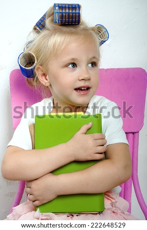 The little girl sits on a bright chair with books in her hands and hair curlers on her head