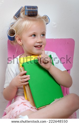The little girl sits on a bright chair with books in her hands and hair curlers on her head
