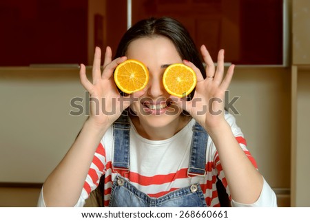 Portrait of a housewife who is holding round orange slices in front of her eyes and smiles. The girl\'s dark hair dressed in blue overalls home.