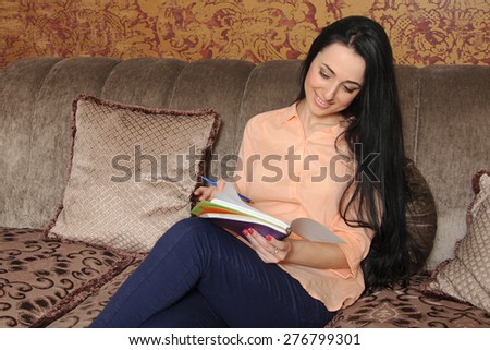 Woman in casual clothes sitting on a couch and makes entries in the diary. She looks down and smiles slightly.