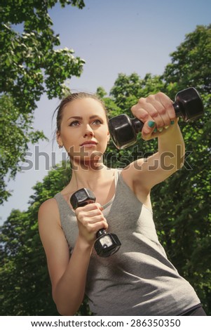 Fitness woman mimics a hit with light dumbbells against a background sky and foliage. The concept of healthy lifestyle, health and beauty