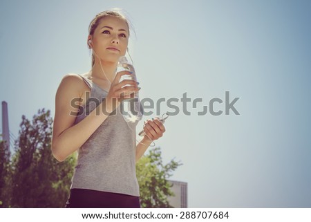 Sport woman drinking water and listening to music with headphones in the phone in the park outdoors