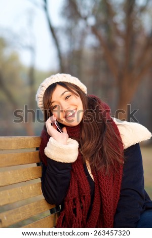 Girl enjoying talking on mobile phone on bench in park in the Winter looking at camera