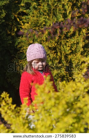 Baby in blue dress and red blouse is surrounded by greenery in the garden