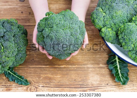 A child holding  broccoli in her hands over a farmhouse table with broccoli in a bowl and on the table ether side of her, room at the bottom for text