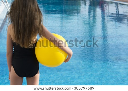 Young child with long brown hair, wearing a black swimming costume stood facing an empty pool, holding a yellow beach ball. room from text and copy space