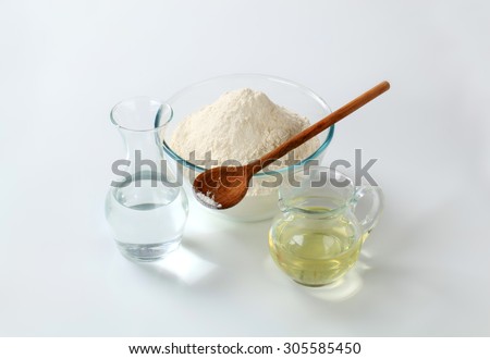 white wheat flour in a glass bowl with a wooden spoon, a carafe of cold water and a jug of sunflower oil