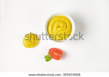 bowl and swirl of yellow mustard and slice of tomato on white background