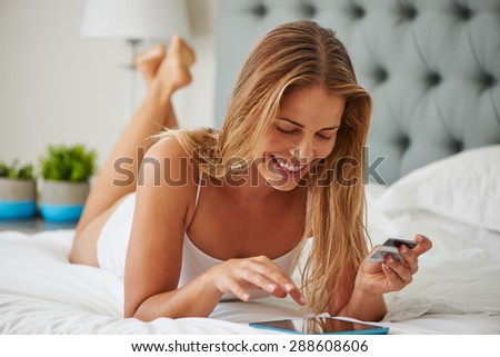 Attractive young woman shopping online using digital tablet lying in bed at upmarket home green plants out of focus in background