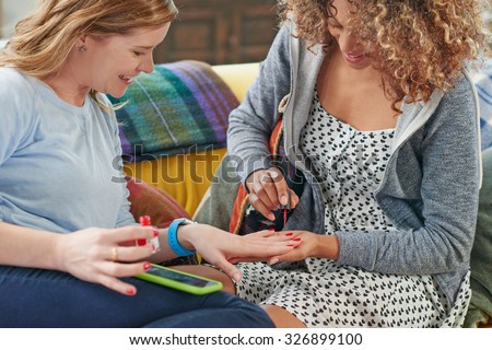 Young mixed race girl painting smiling older womans finger nails red sitting on sofa resting mobile phone on lap perching on cushions