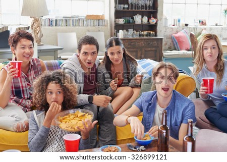 Multi racial group of  student friends sports fans excited drinking beer eating chips watching football on TV