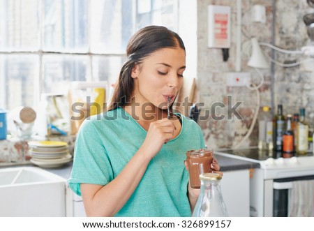 Pretty hispanic girl  cheeky face eating chocolate spread from  jar using spoon wearing pajamas at home by window