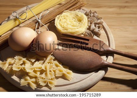Composition with different kinds of pasta, eggs and wooden kitchen set placed on round cutting board
