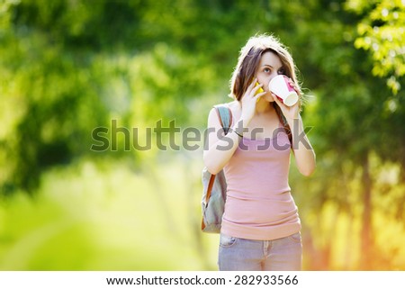 Woman wearing casual clothes talking on phone and drinking beverage in disposable coffee cup. Blonde woman with smartphone and coffee in summer day park.
