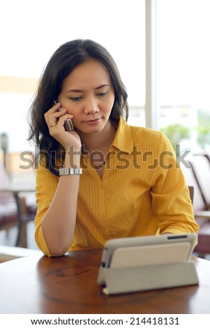 Young Pretty Asian Woman Talking On Phone And Enjoying Touching Tablet In Cafe