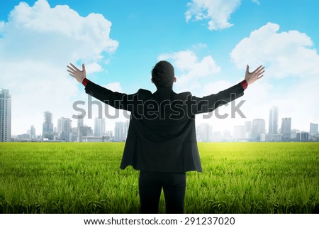 Asian business man standing on green meadow facing city. Business vision  concept