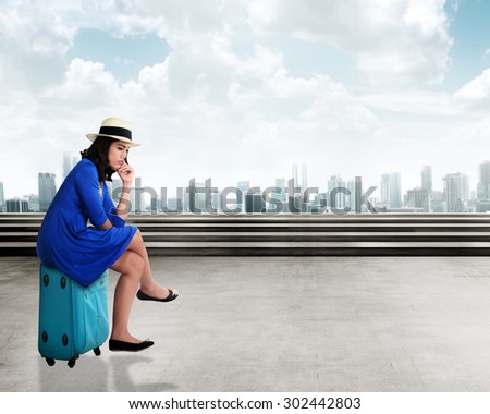 Asian young woman traveler with sad face, sitting on the suitcase over city background