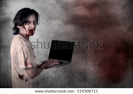 Scary male zombie typing with laptop over grunge background