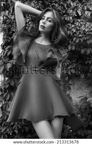 Beautiful sexy woman brunette model in a short blue silk fashionable stylish dress long beautiful tanned legs in high heels on a background of green foliage of summer nature black and white