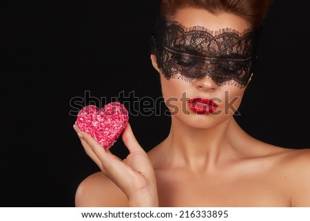 Young beautiful sexy woman with dark lace on eyes bare shoulders and neck, holding cake shape of heart to enjoy the taste and are dieting, feeling temptation, teeth passion sex red lips