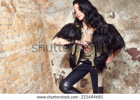 Beautiful sexy woman long brunette hair in black fur and Leather trousers near yellow and red brick wall on background in rock style