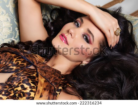 Beautiful sexy brunette woman with wavy hair in a bright multi-colored suit wild animal dress with rings bracelets accessories, or evening hairstyle and makeup lying on the bed with cushions