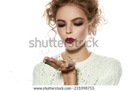 Beautiful sexy young blond girl with wavy hair bright evening make-up red lips long fluffy eyelashes holds palm with gold sequins in front of him and blows them a holiday New Year Christmas joy fun