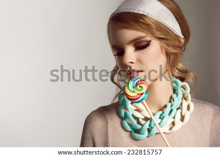 Beautiful sexy young girl with wavy blond hair and silk scarves, soft natural make-up in a shiny silver jacket, blue and white necklace around the neck in the form of a chain, candy on a stick caramel