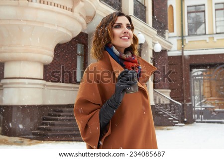 Beautiful sexy young woman with curly brown hair with bright makeup wearing brown coat goes on snow-covered street past shops with a cup of hot coffee, cappuccino, tea, Christmas and New Year Winter