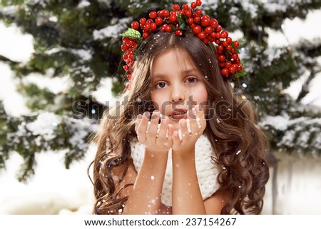 Beautiful little girl with long hair in winter white sweater with a wreath of red berries on a head sits in the snow under the Christmas tree gift for New Year Merry Christmas card