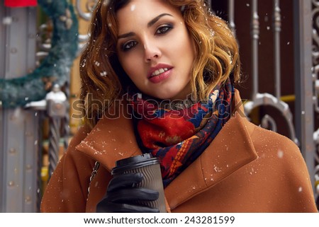 Beautiful sexy young woman with curly brown hair with bright makeup wearing brown coat goes on snow-covered street past shops with a cup of hot coffee, cappuccino, tea, Christmas and New Year Winter