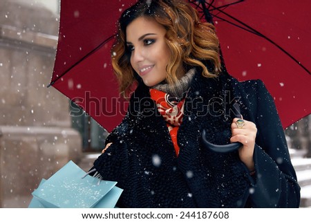 Beautiful sexy young woman with curly brown hair with bright makeup wearing a black coat walking on snow-covered streets past shops with red umbrella and gift packs for Christmas and New Year Winter