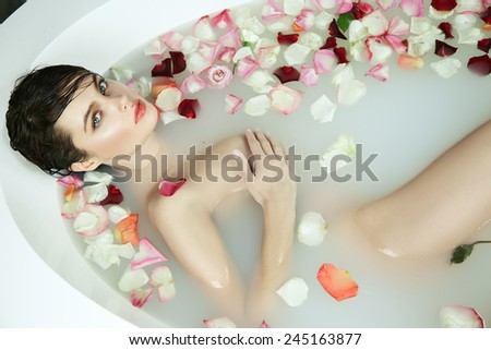 Beautiful young sexy girl with dark hair wet, evening makeup, takes bath with milk and red pink roses petals and candles in romantic atmosphere, beauty cosmetic salon spa for woman at Valentine\'s Day