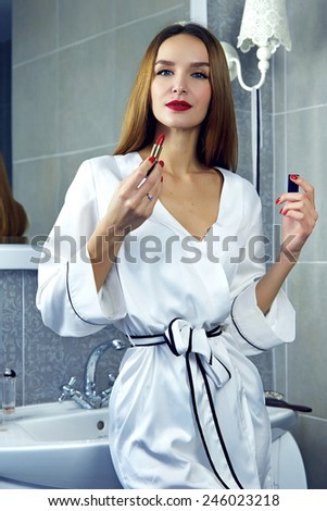 Beautiful young sexy woman with dark hair and evening make-up red lips wearing a white silk robe color lipstick standing in the bathroom at the mirror with makeup and perfume