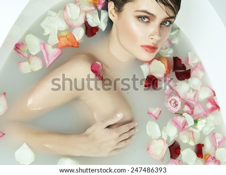 Beautiful young sexy girl with dark hair wet, evening makeup, takes bath with milk and red pink roses petals and candles in romantic atmosphere, beauty cosmetic salon spa for woman at Valentine\'s Day