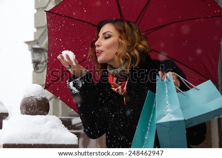 Beautiful sexy young woman with curly brown hair with bright makeup wearing a black coat walking on snow-covered streets past shops with red umbrella and gift packs for Christmas and New Year Winter
