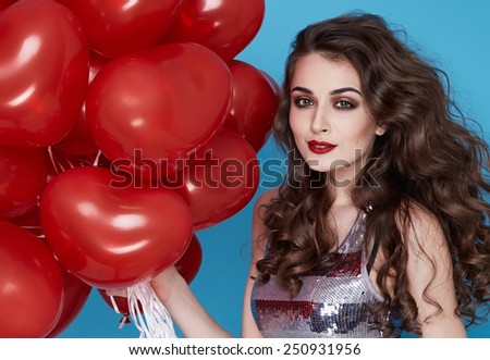 Beautiful young sexy girl with long hair brunette woman with evening make-up cosmetics dressed in a silver dress holding in hand many red balloons party Valentine's Day heart and love
