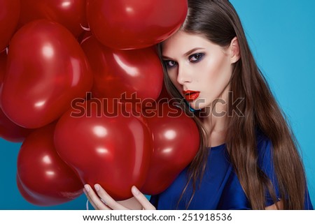 Beautiful young sexy girl with long hair brunette woman with evening make-up cosmetics dressed in a blue silk dress in hand many red balloons party Valentine\'s Day heart and love