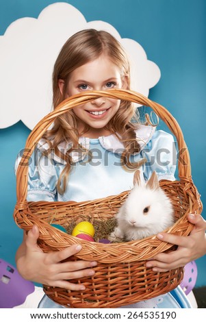 Little fluffy white Easter bunny sitting in a wicker basket on a background of blue sky and white clouds small beautiful cute  blonde girl holding