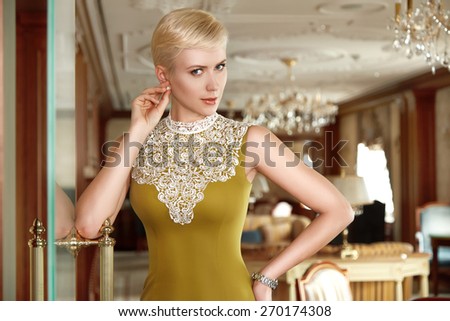 Beautiful young sexy blonde with short hair exquisite evening make-up sitting in a room with an interior in fashionable stylish dress bare shoulders lamp sofa Baroque