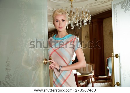 Beautiful young sexy blonde with short hair exquisite evening make-up standing at the door an interior in fashionable stylish dress bare shoulders lamp sofa Baroque