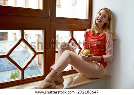 Beautiful young sexy blond woman with makeup business woman seating near big window rest in room in the luxurious interior in stylish clothes collection holding green apple smart looking perfect smile