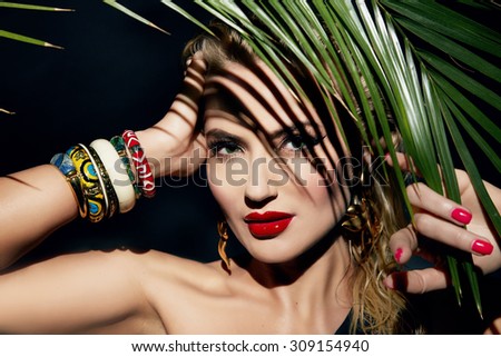 Beautiful young sexy young woman model with perfect figure and tanned skin body bright makeup oil and water drops on the hand wearing lots of bracelet palm leaves in the jungle shadows shining sun