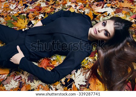 Beautiful young sexy brunette girl with long silky hair evening makeup perfect figure dressed stylish fashion black silk pants suit blouse lying golden autumn leaves collection catalog cosmetic fall