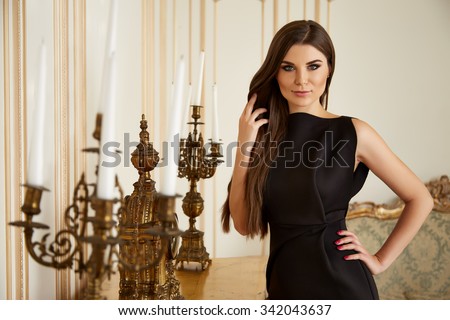 Beautiful sexy young brunette woman with long hair thin slender figure perfect body and pretty face make-up wearing black short silk dress small bag high heels gold interior luxury furniture party
