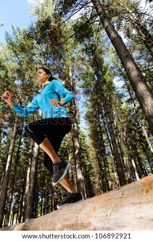 Woman trail running in the woods and jumping over logs while on extreme outdoor fitness training in forest.
